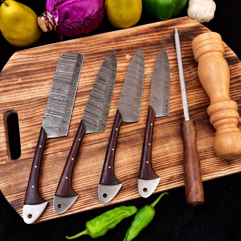May include: Five Damascus steel kitchen knives with wooden handles on a wooden cutting board. The knives have a hammered finish and are arranged in a row. There is a wooden pepper mill and a metal sharpening steel on the board.