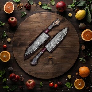May include: Two kitchen knives with wooden handles and patterned blades are displayed on a wooden cutting board surrounded by fruit and greenery.