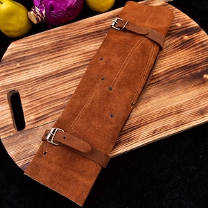 May include: A brown leather knife roll with three straps and buckles. The roll is lying on a wooden cutting board.