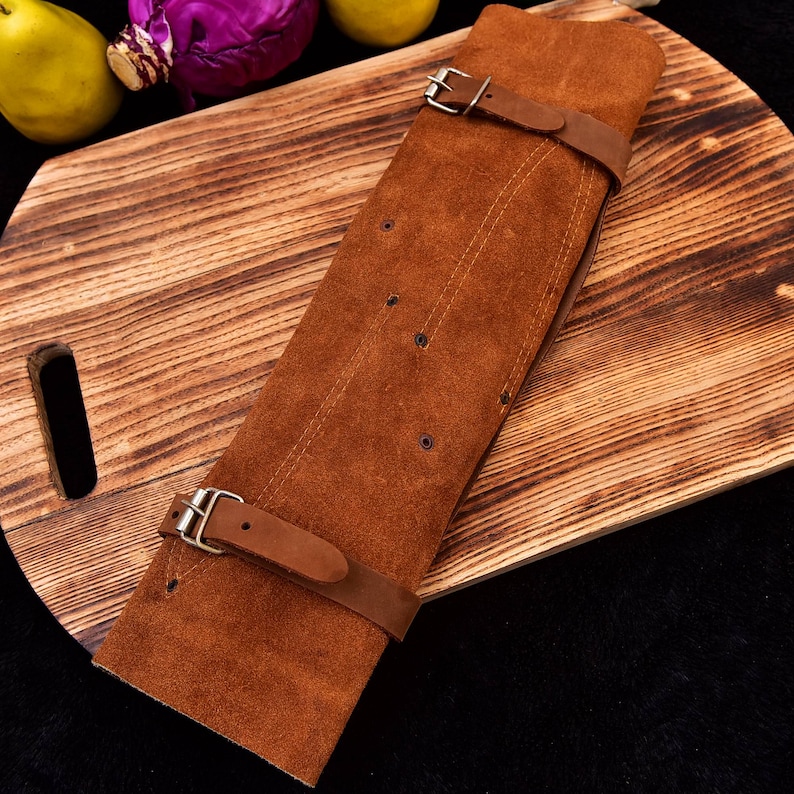 May include: A brown leather knife roll with three straps and buckles. The roll is lying on a wooden cutting board.
