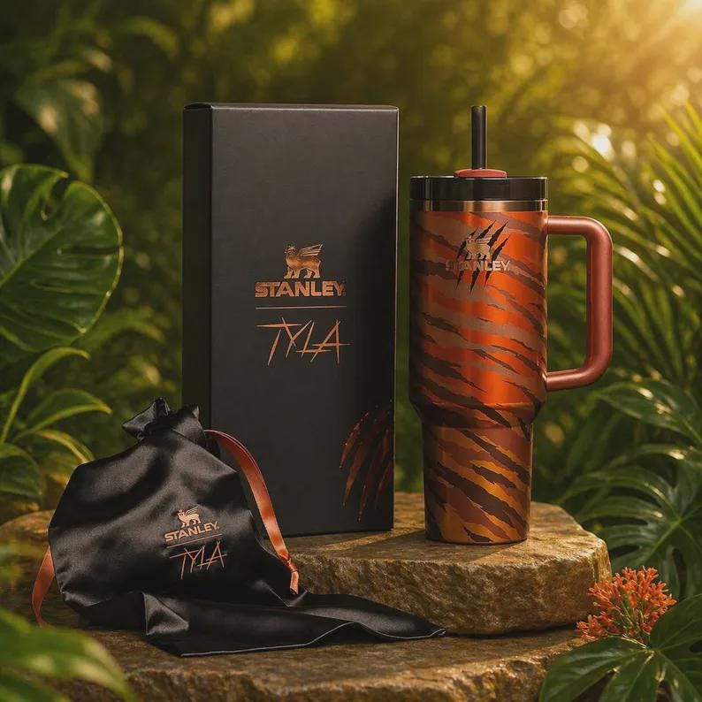 K&ouml;nnte beinhalten: Ein Stanley Tyla Becher mit Tigerstreifen-Design, eine schwarze Box und ein schwarzer Satinbeutel. Der Becher hat einen Griff und einen Strohhalm. Die Box und die Tasche tragen das Stanley-Logo und den Namen "Tyla" in Gold.