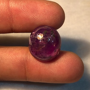 May include: A round, polished cabochon gemstone with a deep, translucent purple hue. The stone features internal inclusions and a smooth, reflective surface that catches the light, displaying variations in color intensity throughout the gem.