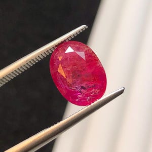 May include: An oval-cut, vibrant red gemstone with a faceted surface, held by metal tweezers. The stone displays internal inclusions and varying shades of deep crimson, reflecting light across its polished, multi-faceted top surface.