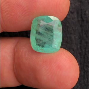 May include: A cushion-cut emerald gemstone with a vibrant green hue and visible internal inclusions. The stone features a faceted surface that reflects light, showcasing a translucent quality and a distinct square shape with rounded corners.
