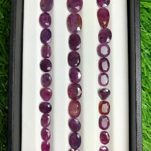 Puede incluir: Una colección de piedras preciosas facetadas de forma ovalada en tonos púrpura y rosa rojizo, dispuestas en tres filas verticales dentro de una bandeja de exhibición negra. Las piedras muestran matices magenta y violeta con superficies reflectantes.