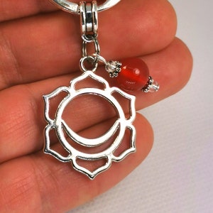 May include: Silver-toned keychain featuring a chakra symbol pendant and a red gemstone bead. The pendant has a floral design with a crescent moon shape in the center. The keychain is attached to a silver ring.