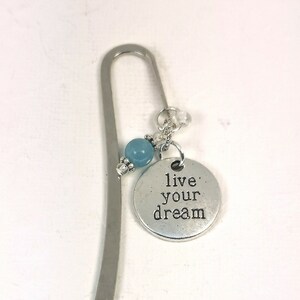 May include: A silver metal bookmark with a curved design. It features a light blue bead and a round charm that reads "live your dream". The bookmark is a simple and elegant accessory.