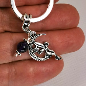 May include: Silver-coloured keyring featuring a crescent moon with a star and a fairy design. A dark purple bead hangs below the moon. The keyring is attached to a silver ring.