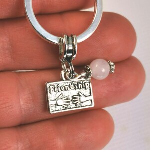 Op de afbeelding: Zilverkleurige sleutelhanger met een rechthoekige bedel met de tekst "Friendship" en twee handen die elkaar vasthouden. Een roze, bolvormige kraal en een zilveren ring maken het ontwerp compleet. De sleutelhanger wordt in een hand gehouden.
