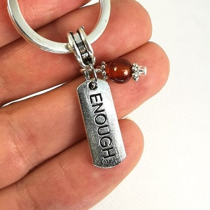 May include: Silver keychain with a round ring and a rectangular charm that reads "ENOUGH". A small, round, reddish-brown bead and decorative silver bead are attached to the charm. The keychain is held in a hand.