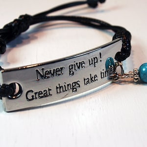 Op de afbeelding: Een zwart koordarmband met een zilveren plaat met de tekst "Never give up! Great things take time." De armband heeft turquoise kralen en zilveren accenten.