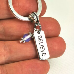 May include: Silver keyring with a rectangular charm inscribed with the word "BELIEVE". A small purple and white bead is attached. The keyring is held in a hand.