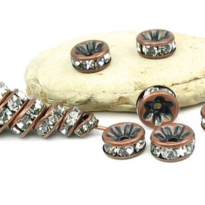 May include: A set of copper-toned metal bead spacers with clear crystal rhinestones. The spacers are round and have a raised center. The spacers are arranged in a line on a light brown rock.