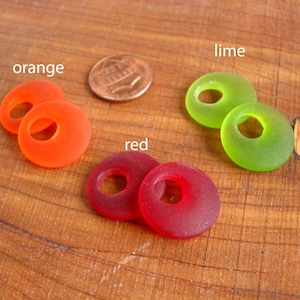 May include: Four glass beads in different colors: orange, red, lime green, and red. The beads are round with a hole in the center. They are arranged in a row on a wooden surface.