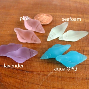 Peut inclure: Six coquillages en verre de différentes couleurs, dont rose, lavande, mousse de mer et aqua. Les coquillages sont disposés sur une surface en bois.