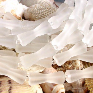 K&ouml;nnte beinhalten: Nahaufnahme einer Ansammlung von gefrosteten Glasperlen in einer hellen, durchscheinenden wei&szlig;en Farbe. Die Perlen sind in Form einer Schleife oder einer Sanduhr geformt, mit einer schmalen Mitte und breiteren Enden. Die Perlen sind in einem zuf&auml;lligen Muster angeordnet, wobei sich einige &uuml;berlappen.