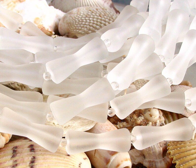 K&ouml;nnte beinhalten: Nahaufnahme einer Ansammlung von gefrosteten Glasperlen in einer hellen, durchscheinenden wei&szlig;en Farbe. Die Perlen sind in Form einer Schleife oder einer Sanduhr geformt, mit einer schmalen Mitte und breiteren Enden. Die Perlen sind in einem zuf&auml;lligen Muster angeordnet, wobei sich einige &uuml;berlappen.