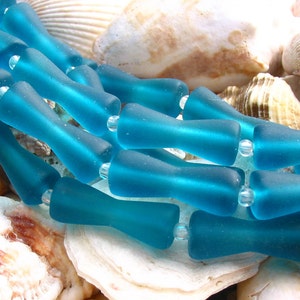 K&ouml;nnte beinhalten: Eine Reihe von t&uuml;rkisfarbenen Glasperlen, jede Perle ist tropfenf&ouml;rmig mit abgerundetem Ende. Die Perlen sind in einer Reihe angeordnet und liegen auf einem Bett aus Muscheln.