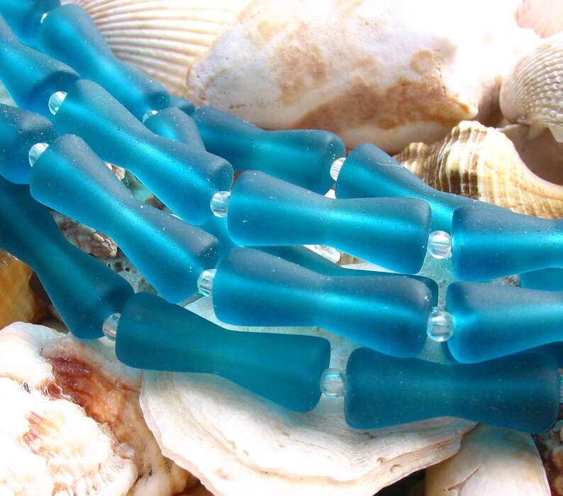 K&ouml;nnte beinhalten: Eine Reihe von t&uuml;rkisfarbenen Glasperlen, jede Perle ist tropfenf&ouml;rmig mit abgerundetem Ende. Die Perlen sind in einer Reihe angeordnet und liegen auf einem Bett aus Muscheln.