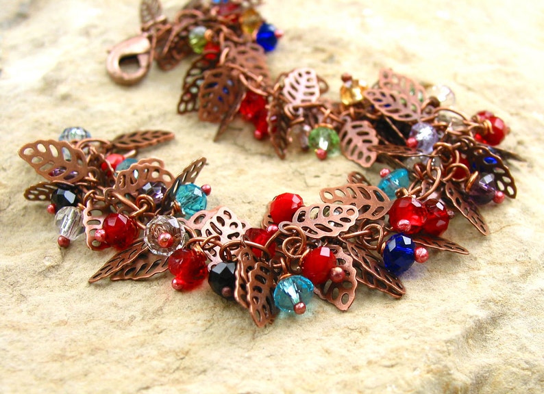 May include: A copper bracelet with a leaf design and colorful beads. The beads are red, blue, green, and clear. The bracelet is made of copper wire and has a delicate, feminine look.