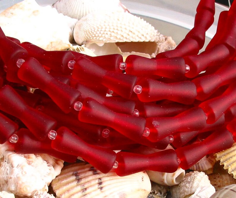 K&ouml;nnte beinhalten: Eine Str&auml;hne roter Glasperlen, jede Perle ist in Form einer kleinen Flasche mit abgerundetem Oberteil und leicht ausgestelltem Boden geformt. Die Perlen sind in einem lockeren Haufen angeordnet, wobei sich einige Perlen &uuml;berlappen.