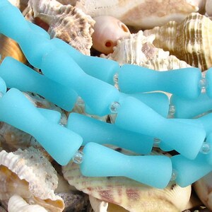 K&ouml;nnte beinhalten: Eine Reihe von hellblauen Glasperlen mit mattem Finish. Die Perlen sind tropfenf&ouml;rmig mit abgerundetem Ende. Die Perlen sind in einer Reihe angeordnet und liegen auf einem Bett aus Muscheln.