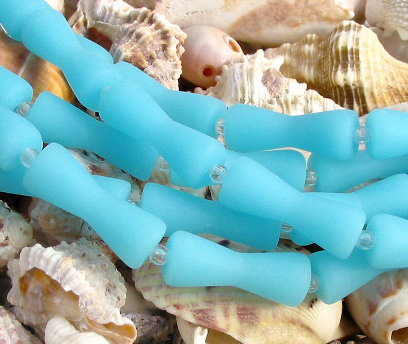 K&ouml;nnte beinhalten: Eine Reihe von hellblauen Glasperlen mit mattem Finish. Die Perlen sind tropfenf&ouml;rmig mit abgerundetem Ende. Die Perlen sind in einer Reihe angeordnet und liegen auf einem Bett aus Muscheln.