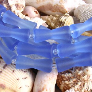 K&ouml;nnte beinhalten: Eine Reihe von hellblauen Glasperlen mit mattem Finish. Die Perlen sind tropfenf&ouml;rmig mit abgerundetem Ende. Die Perlen sind in einer Reihe angeordnet und liegen auf einem Bett aus Muscheln.