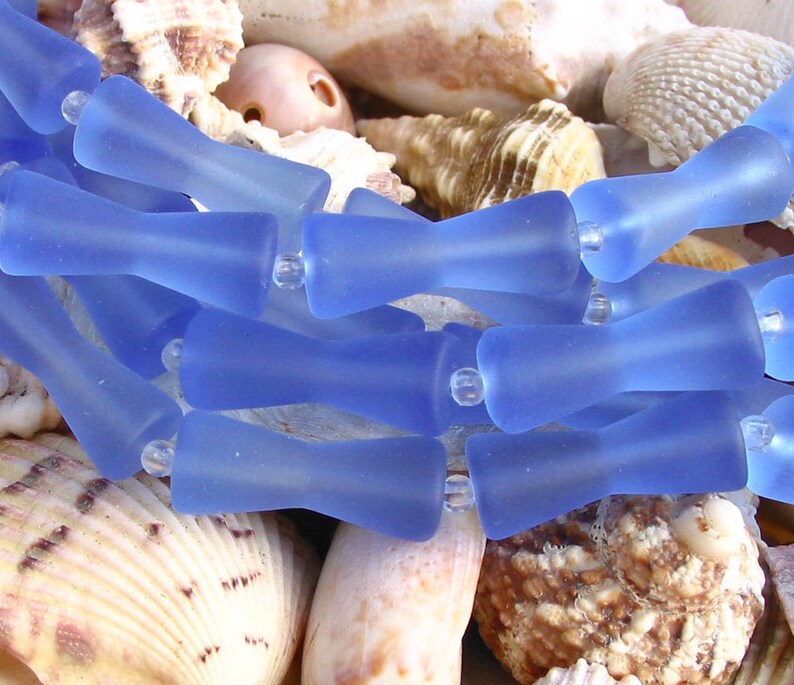 K&ouml;nnte beinhalten: Eine Reihe von hellblauen Glasperlen mit mattem Finish. Die Perlen sind tropfenf&ouml;rmig mit abgerundetem Ende. Die Perlen sind in einer Reihe angeordnet und liegen auf einem Bett aus Muscheln.