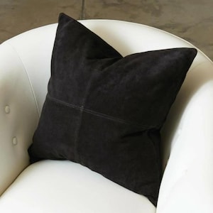 May include: A dark charcoal-coloured square cushion with a stitched cross design. The cushion is made of a soft, suede-like material and is placed on a white chair. This decorative cushion adds a touch of elegance to any living space.