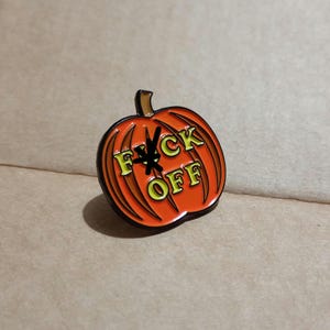 May include: An orange pumpkin-shaped enamel pin with black outlines and a brown stem. The centre of the pumpkin features the text FUCK OFF in yellow lettering, with a black star symbol replacing the letter U in the word FUCK.