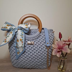 Op de afbeelding: Lichtblauwe gehaakte handtas met een houten handvat en een lichtblauwe schouderriem. Een gouden ketting is aan de tas bevestigd. Een bloemen sjaal is aan het handvat gebonden. Een kleine vaas met roze bloemen staat rechts.