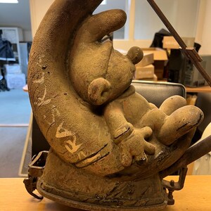 May include: A rusty metal sculpture of a cartoon character sitting on a crescent moon. The sculpture is painted in shades of brown and white.