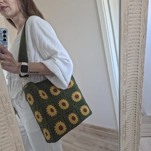 May include: A green crochet tote bag with a sunflower design. The bag has a long, olive green strap and gold-coloured hardware. The sunflower pattern is made with yellow and brown yarn.