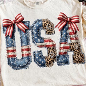 May include: Cream-colored t-shirt featuring the word "USA" in a patriotic design. The letters are a patchwork of denim, American flag stripes, and leopard print, adorned with red and white striped bows. The shirt is a digital download.