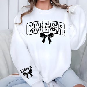 May include: White sweatshirt with the words "CHEER mom" in black block letters, a black bow graphic, and the name "EMMA" with a bow on the sleeve. The sweatshirt is a casual style.