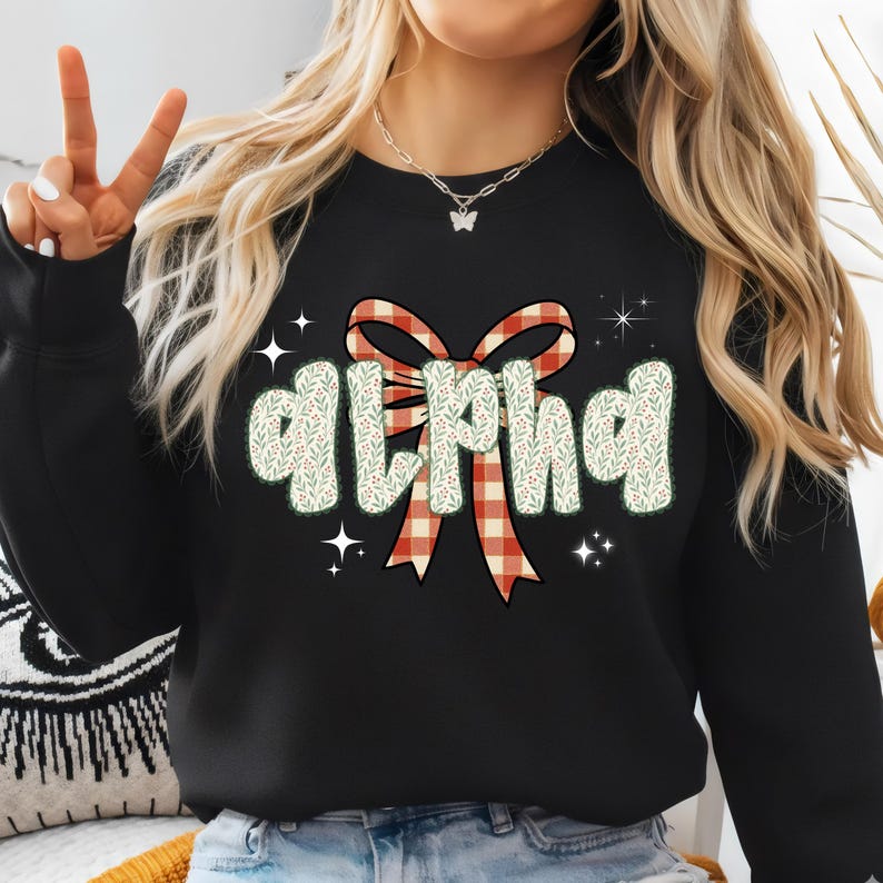 Pode incluir: Camisola preta com a palavra "ALPHA" em uma fonte decorativa. As letras s&atilde;o preenchidas com um padr&atilde;o floral verde e branco, e a palavra &eacute; encimada por um la&ccedil;o xadrez vermelho e branco. Explos&otilde;es de estrelas brancas s&atilde;o espalhadas ao redor da palavra.