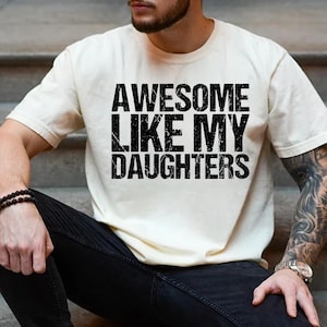 Op de afbeelding: Cr&egrave;mekleurig T-shirt met de tekst "AWESOME LIKE MY DAUGHTERS" in vette, zwarte letters. Het shirt heeft korte mouwen en lijkt van zacht materiaal te zijn. De tekst heeft een versleten uitstraling.