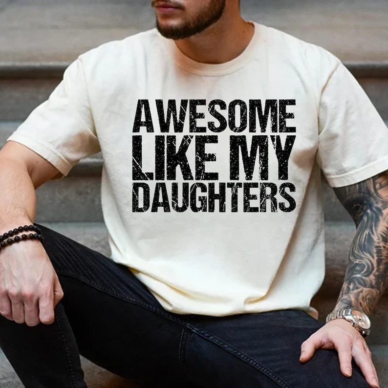 Op de afbeelding: Cr&egrave;mekleurig T-shirt met de tekst "AWESOME LIKE MY DAUGHTERS" in vette, zwarte letters. Het shirt heeft korte mouwen en lijkt van zacht materiaal te zijn. De tekst heeft een versleten uitstraling.