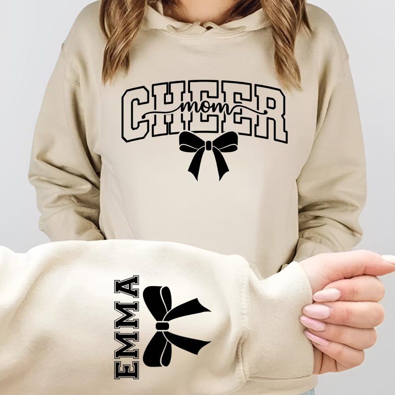 May include: A beige hoodie with the words "CHEER mom" in black block letters and a black bow graphic. The sleeve features the name "EMMA" in black block letters with a black bow graphic.