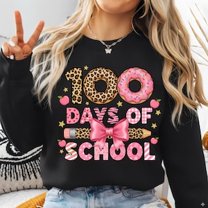 Puede incluir: Sudadera negra con un gráfico colorido que celebra los 100 días de escuela. El diseño incluye una rosquilla con estampado de leopardo, una rosquilla rosa glaseada, un lazo rosa y las palabras "100 Days of School".