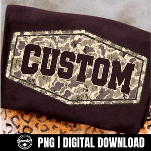 May include: A dark brown t-shirt featuring a central hexagonal graphic with a distressed camouflage pattern in shades of green, tan, and brown. The word CUSTOM is printed in large, bold, dark brown block letters across the center.