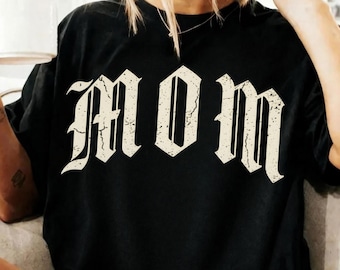 Può includere: T-shirt nero con la scritta "MOM" in un carattere gotico invecchiato. Il testo color crema contrasta con il tessuto scuro. La maglietta ha una vestibilità ampia.