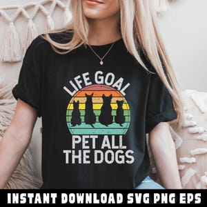 May include: Black t-shirt with the text "LIFE GOAL" above a graphic of four dog silhouettes inside a sunset-colored circle. Below the circle, the text reads "PET ALL THE DOGS". The shirt is a casual style.