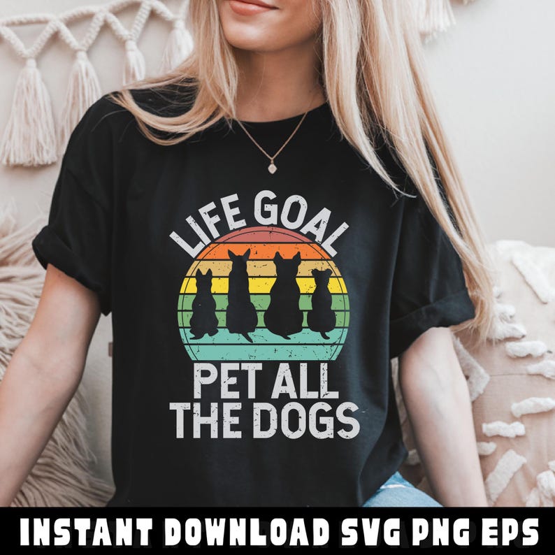 May include: Black t-shirt with the text "LIFE GOAL" above a graphic of four dog silhouettes inside a sunset-colored circle. Below the circle, the text reads "PET ALL THE DOGS". The shirt is a casual style.