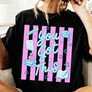 Peut inclure: T-shirt noir avec un motif à rayures roses et blanches. Le t-shirt présente le texte "You Got This" en écriture bleu clair, ainsi que des illustrations d'objets scolaires tels qu'un crayon, un chapeau de remise de diplôme et une pomme.