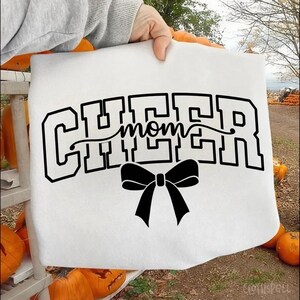 May include: White sweatshirt with the words "CHEER mom" in black block letters and a black bow. The sweatshirt is held up by a person's hand. Pumpkins are in the background.