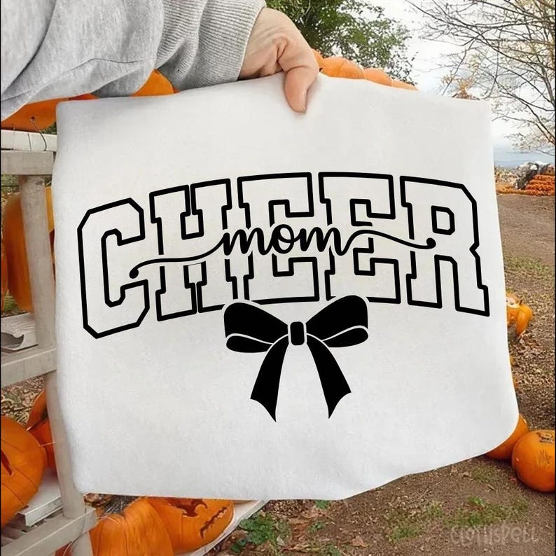 May include: White sweatshirt with the words "CHEER mom" in black block letters and a black bow. The sweatshirt is held up by a person's hand. Pumpkins are in the background.