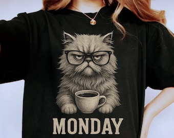 May include: Black t-shirt featuring a grumpy cat wearing glasses and holding a coffee cup. The word "MONDAY" is printed below the cat. The shirt design is a digital illustration, perfect for cat lovers.