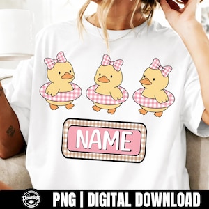 Könnte beinhalten: Weißes T-Shirt mit drei Cartoon-Entenküken, die rosa Vichy-Schwimmreifen und Schleifen tragen. Darunter ein rosa Rechteck mit dem Text "NAME", umrandet von einem braunen Vichy-Muster. Digitaler PNG-Download.